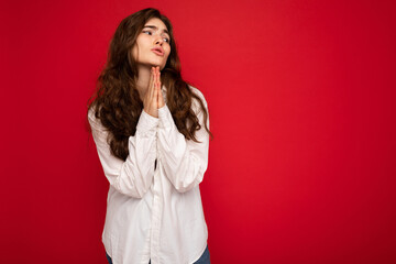 Portrait of beautiful young curly brunette woman wearing white shirt isolated on red background with copy space, praying and dreaming with sincere emotions