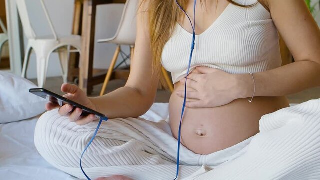 Young pregnant woman sits on the bed with a smartphone in headphones. She actively communicates online - write and send messages via her smartphone.