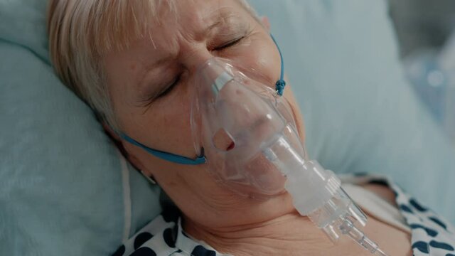 Close up of elder patient with oxygen mask laying in bed. Sick woman with respiratory issues breathing heavily and hyperventilating, using tube to cure coronavirus symptoms during pandemic