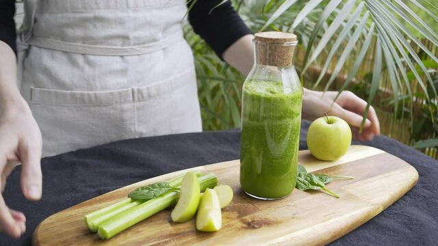 Woman puts wooden tray with glass bottle of smoothie apples and celery stems on table covered with black fabric near lush tropical plants close view