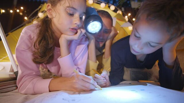 Brother and sister draw while daddy holds glowing flashlight spending time together by play tent with fairy lights in cottage yard in evening closeup