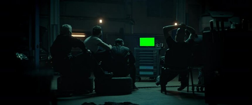 WIDE Car garage workshop workers in uniforms watching portable TV in the evening, sport event, celebrating a goal. Green screen, chroma key. Shot with 2x anamorphic lens