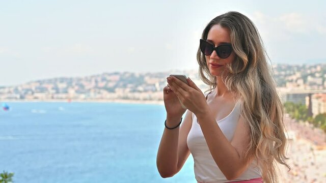 Caucasian smiling woman in sunglasses using smartphone with view of Nice on the background, France