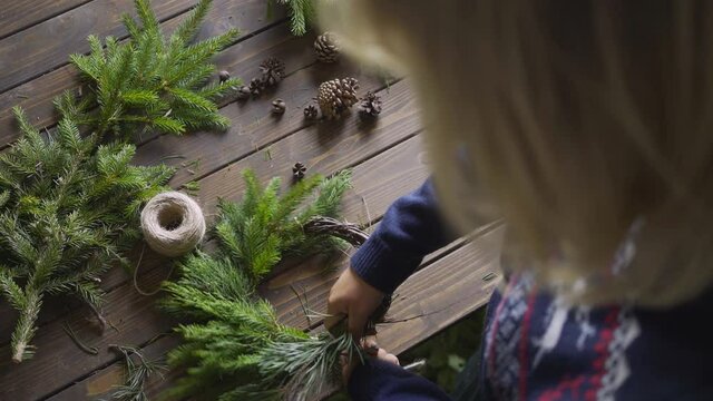 Aged wooden table in authentic florist workshop. Backside view female arranges and attache evergreen fir branches with rope, preparing handmade xmas wreath. DIY and handcraft concept