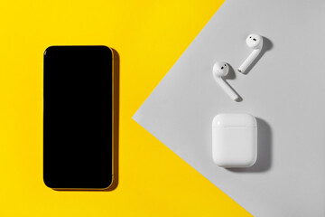 Smartphone and wireless headphones on yellow and gray background.