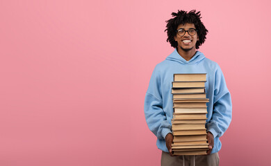 Back to school concept. Happy black teen guy holding huge stack of books, smiling at camera on pink...