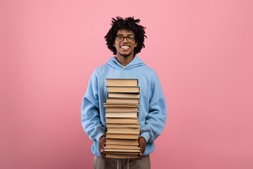 Cheerful black teenager standing with big stack of books, having lots of homework on pink studio...