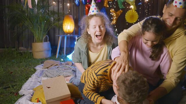 Joyful little son puts face into and bites delicious cake celebrating birthday with parents and sister in decorated suburban cottage yard in evening