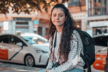 Mujer joven fotógrafa con su cámara de fotos en la ciudad tomándose un descanso sentada en un banco junto a los taxis y el transporte publico para continuar con su ruta de viaje por España