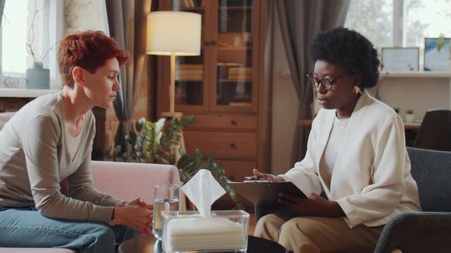 African American psychologist holding clipboard and talking with Caucasian woman while giving her consultation in office