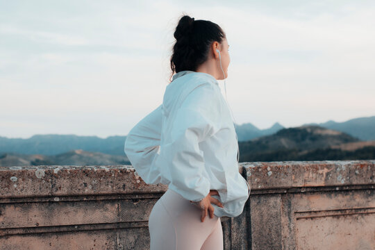 Muchacha joven de cabello oscuro moreno observando las monta&ntilde;as detr&aacute;s del muro del pantano flexionando las caderas para hacer ejercicio cardiovascular y recorrer en ruta el pueblo 