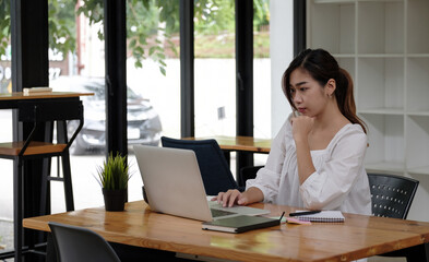 Focused asian business woman working studying online in office looking at laptop, serious employee or student watching webinar writing information in notebook sitting at desk