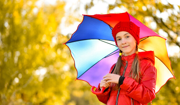 A smiling girl in a red hat and a red jacket walks with a rainbow umbrella in nature. yellow leaves in the background. Autumn. Atmosphere