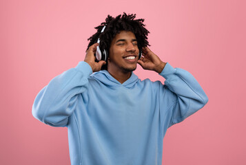 Attractive black teen guy in casual wear listening to music in headphones, closing eyes and relaxing on pink background