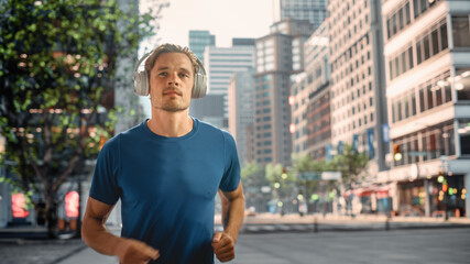 Handsome Young Man is Jogging on a Street of a Big City Center. Male is Running in Blue T-Shirt, Wearing Wireless Headphones with His Favourite Music Playing. Urban Training Workout in the Morning.