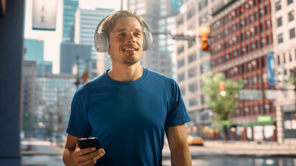 Handsome Young Man Walking on a Street of a Big City Center After Intense Fitness Workout and Using His Smartphone while Listening to Music on Hids Headphones. Urban Training Workout in the Morning.