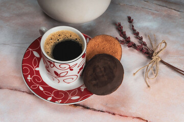 Taza blanca y roja de café negro, con dos galletas de chocolate y un delicado bouquet de lavanda roja atada con cuerda natural, sobre una mesa de piedra de mármol rosa.