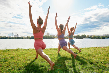 Three fit young beautiful women doing Reverse Warrior Pose, Crescent lunge variation, Viparita Virabhadrasana, working out in park on summer day, wearing sportswear tops, full length. Fitness, sport
