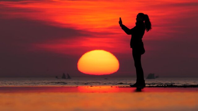 Cheerful woman wave hand to smartphone and say good bye, finish video call and go away. Full length silhouette of lady against sunset at cold autumn beach. Telephoto perspective, large sun move down