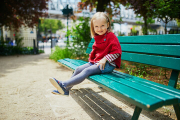 Happy cheerful toddler girl sitting on the bench on a street of Paris