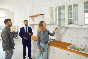 Future tenants or first time buyers meeting real estate agent and looking around house. Boyfriend and girlfriend buying new home. Husband and wife looking at modern white wooden cupboards in kitchen - obrazy, fototapety, plakaty