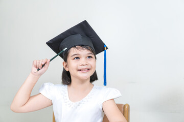 Portrait of cheerful Asian little girl student wear a mortarboard or graduation hat hold pencil and write on notebook with white background with copy space. Education Graduation Concept Stock Photo