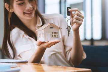 Asian businesswoman or real estate agent holding a sample of a house with keys handing it to a customer at the office.