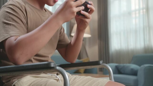 Close Up Excited Young Asian Boy Sitting On Wheelchair Playing Video Game On Mobile Phone And Celebrating In House
