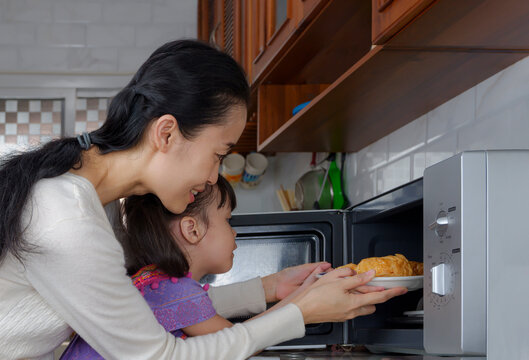 Asian Mother Is Teaching Her Little Daughter To Use A Microwave Oven To Warming Croissants In The Kitchen At Home