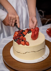 Newlyweds are cutting a beautiful white cake with berries and heart-shaped decor. Close-up
