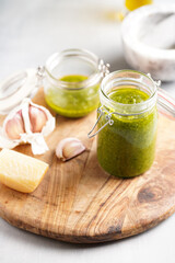 Several small glass mason jars with green italian basil sauce pesto on a round wooden tray with hard cheese pecorino and garlic cloves