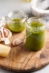 Several small glass mason jars with green italian basil sauce pesto on a round wooden tray with hard cheese pecorino and garlic cloves