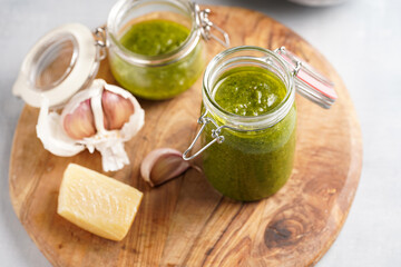 Several small glass mason jars with green italian basil sauce pesto on a round wooden tray with hard cheese pecorino and garlic cloves