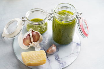 Several small glass mason jars with green italian basil sauce pesto on a round marble tray with hard cheese pecorino and garlic cloves