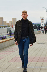 A young man in a black jacket walks along the city embankment. Lighthouse in the background in blur. Acne on the face. Selective focus. Blagoveshchensk, Russia.