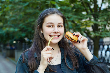 young brunette holds a hamburger in her hands and bites French fries