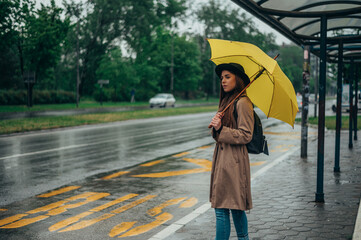 Young beautiful woman holding a yellow umbrella and standing on a bus stop