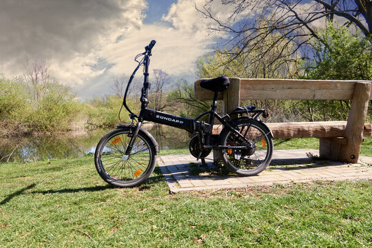 Z&uuml;ndapp electric folding bike leaning against a massive rustic wooden bench in front of a river under sensible cloudy sky in Gifhorn, Germany, April 18, 2020
