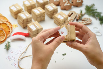 Fototapeta premium Female hands packages gifts for advent calendar. Preparing for Christmas with gingerbread cookies, dried oranges, sprigs of fir. Eco friendly new year holidays