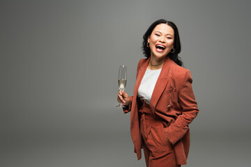 Excited asian woman in jacket holding champagne and looking at camera isolated on grey