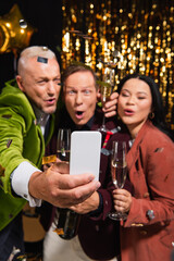 Smartphone in hand of blurred man taking selfie with interracial friends and champagne during new year party on black background