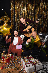 Positive interracial friends with champagne, acoustic guitar and balloons celebrating new year on black background