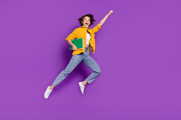 Fototapeta na wymiar Photo of excited funny young woman dressed yellow shirt jumping high holding copybooks rising fist isolated purple color background
