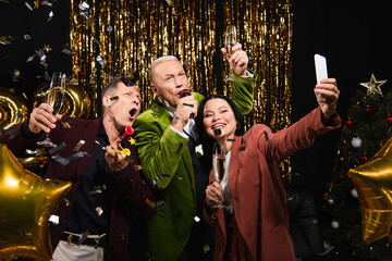 Excited interracial friends taking selfie and singing karaoke during new year party on black...