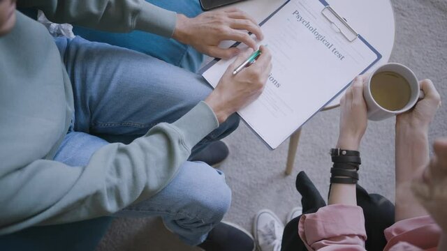 Male patient fills psychological test form talking to specialist with cup of coffee at small table in light office at appointment close upper view