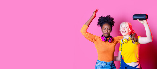 Young diverse black and caucasian women indoor holding wireless bluetooth speaker dancing together...