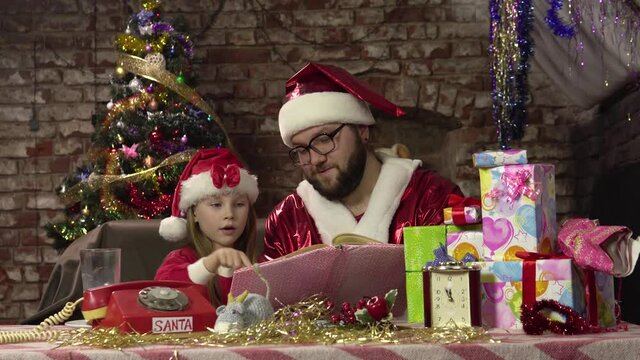Santa Claus sits on armchair and reads book with fairy tales for enthusiastic little girl.4k