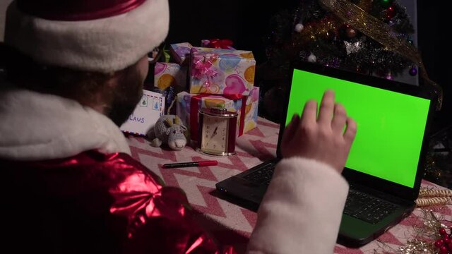 Santa Claus sits at a table with a laptop, waves his hand, congratulates.Green laptop screen.