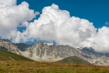 A panoramic view on high Caucasus mountains in Georgia. There are high glaciers in the back. Thick clouds above the sharp peaks. Lush pastures on the sides. Barren peaks.  Idyllic landscape.