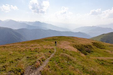 A woman hiking along a narrow pathway in high Caucasus mountains in Georgia. There are high glaciers in the back. Thick clouds above the sharp peaks. Lush pastures on the sides. Barren peaks.